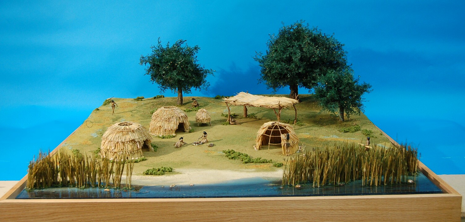 Mission San Miguel village diorama with thatched huts, miniature figures, trees and reeds beside water.